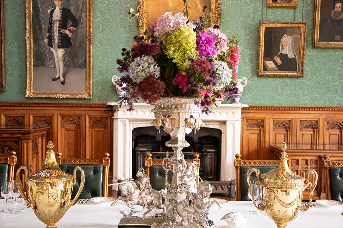 A dining room with paintings hanging on the wall with an elaborate arrangement filled with fresh flowers sitting on top of a silver stand.