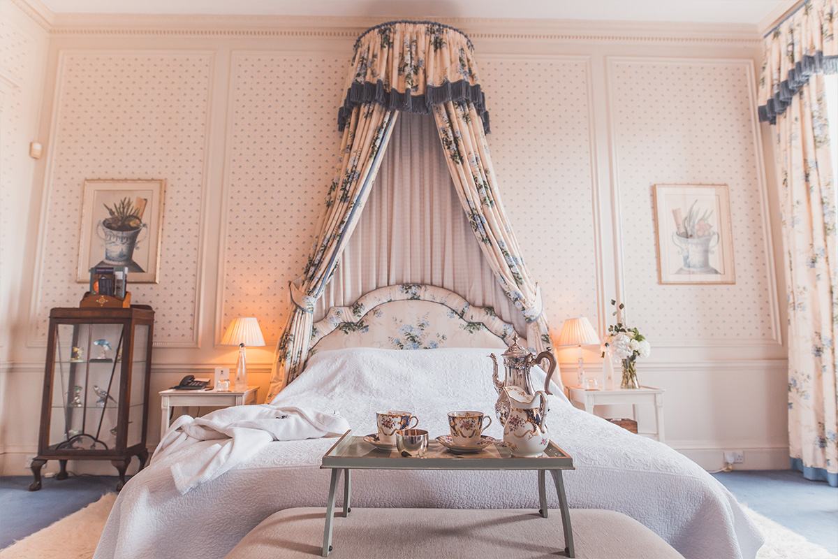 Pastel pink bed with patterned throw, fine china tea set rested on top all underneath a floral canopy. Polka dotted pink wallpaper surrounding