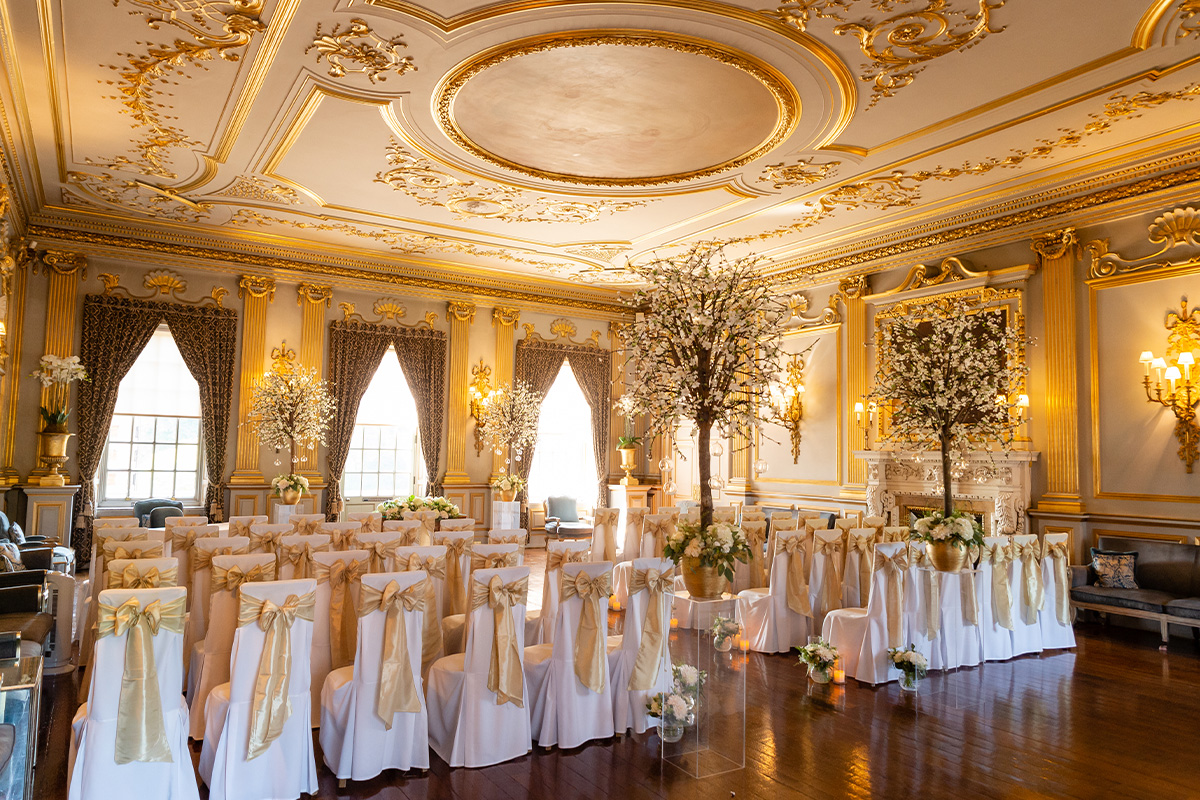 White chairs with golden ribbon draped down the back in a golden embellished room with white trees. Dressed for a wedding ceremony at Knowsley Hall.
