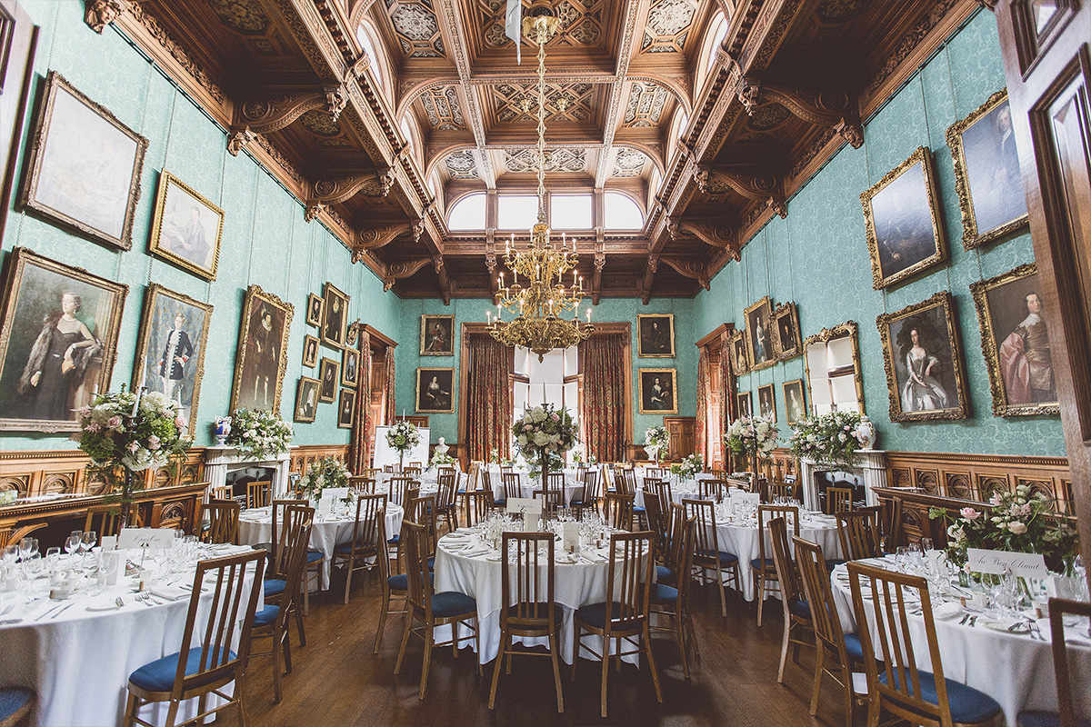 A very grand dining hall with high ceilings with large gold chandelier, paintings handing on the walls, round tables and chars and fresh flowers on each table