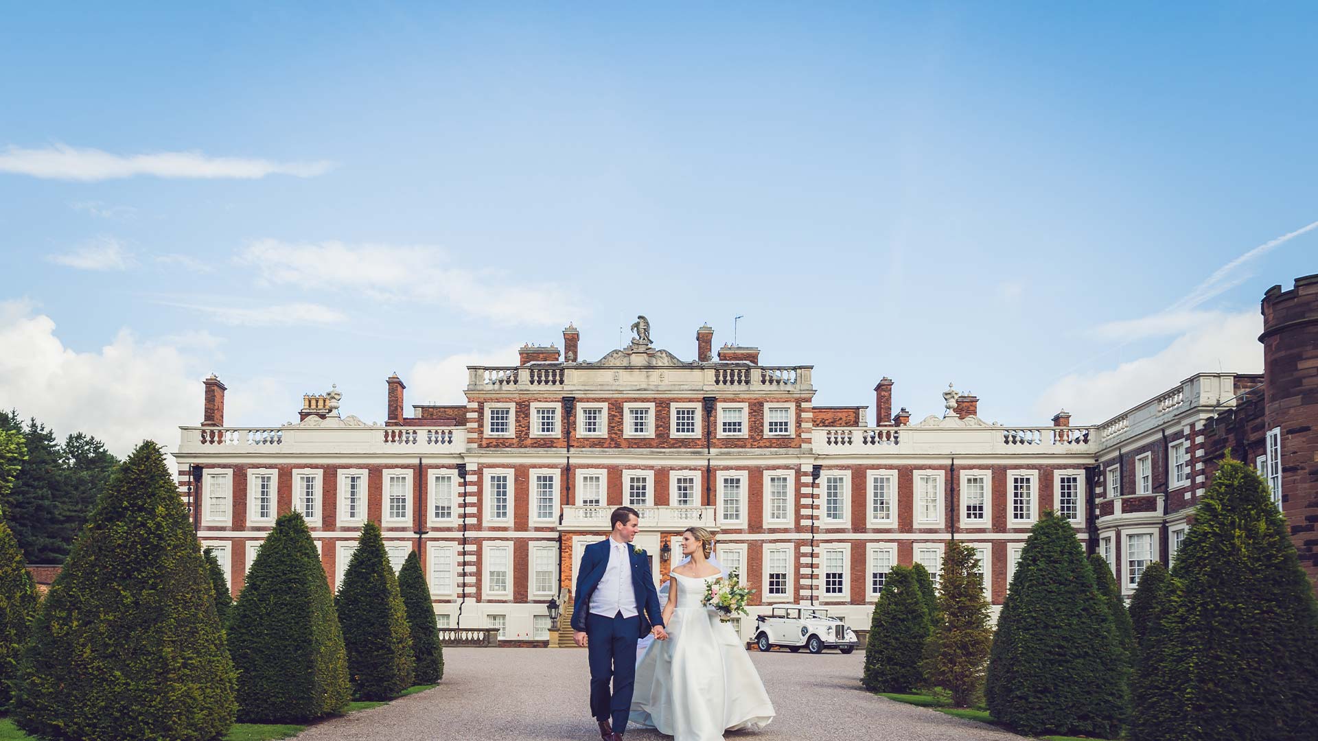 Newlyweds Bride and Groom walking hand in hand outside the front of a mansion house on their summer wedding day