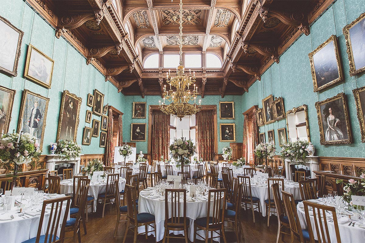 A huge dining room with gold chandelier filled with round tables dressed with white linen, chairs and flowers for a summer wedding